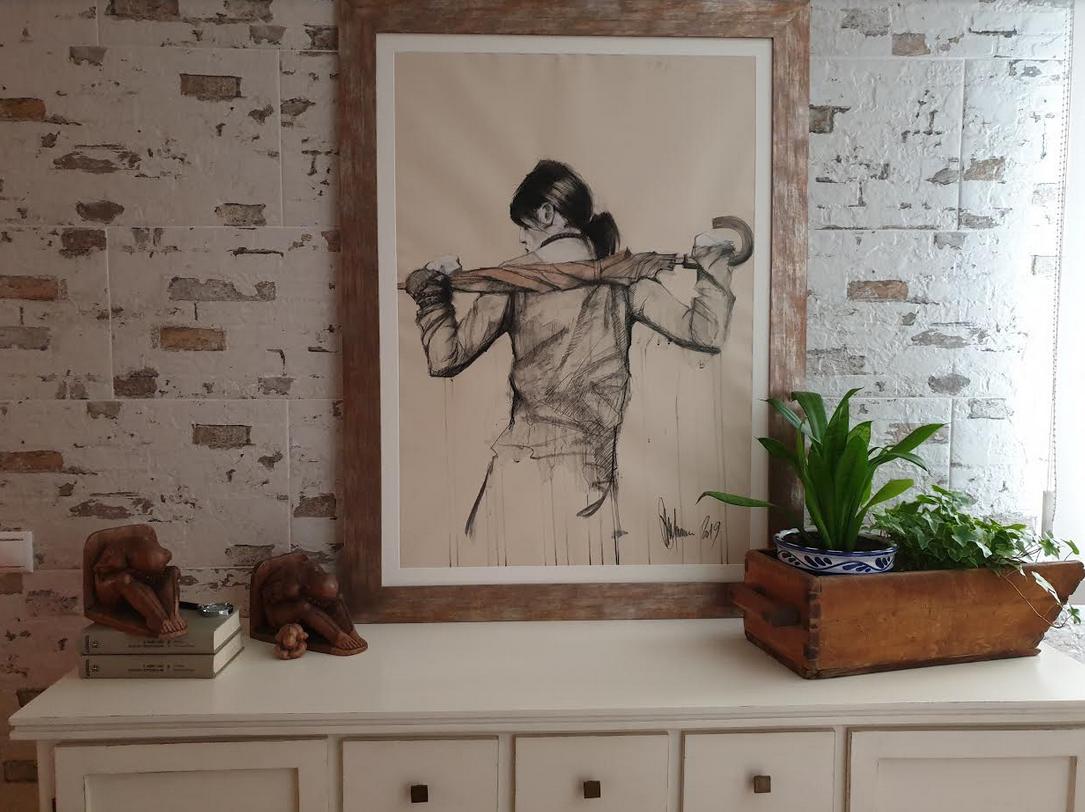Igor Shulman's "Umbrella" artwork in a client's interior space A framed mixed-media drawing of a woman holding an umbrella on her shoulders by Igor Shulman, displayed on a wall above a white cabinet in a client's living room.