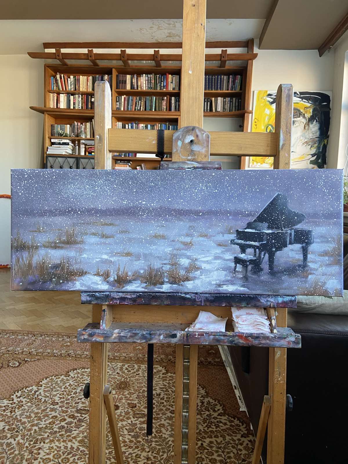 Finished painting of an old grand piano in a snowy field with heavy cloudy sky, displayed on an easel in Igor Shulman's studio.
